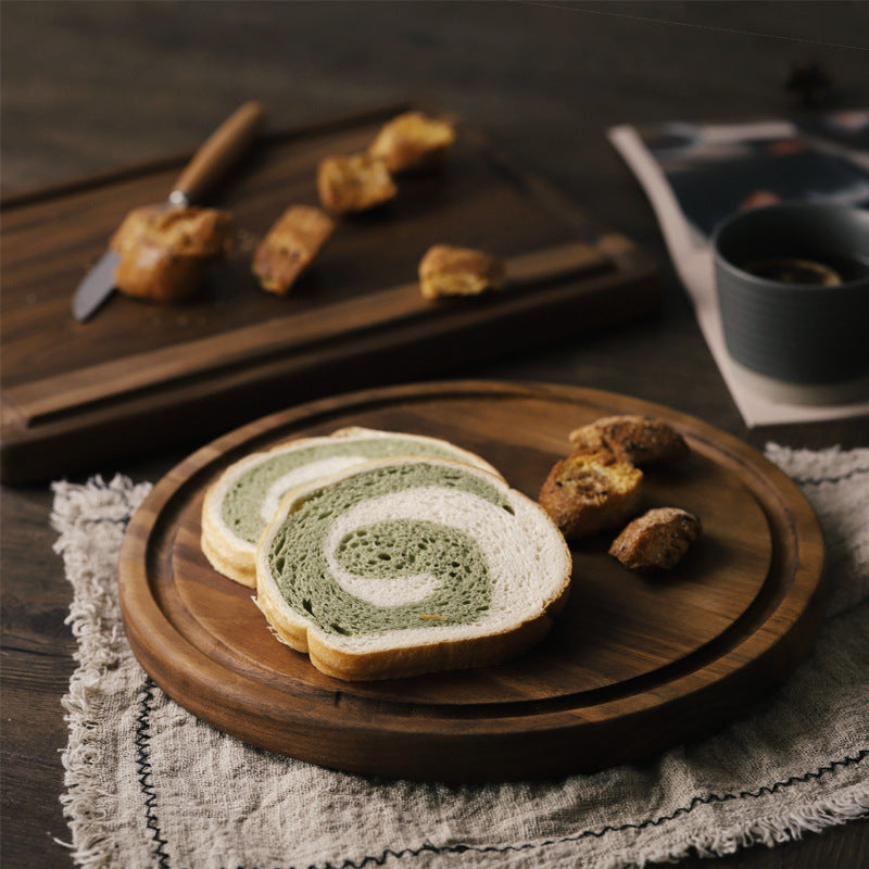 Slices of green-swirl bread on a wooden plate with small pastries on a dark surface.