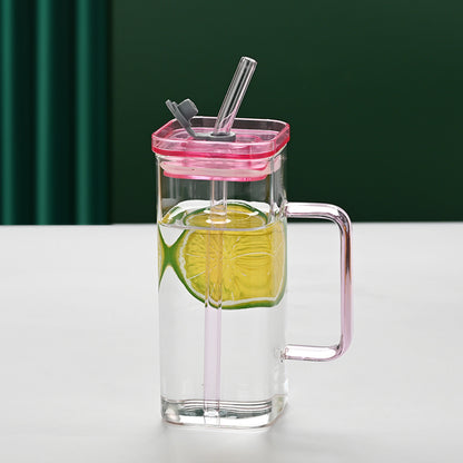 Clear glass mug with pink lid and handle, filled with a green drink and lime slice, on a white surface with green curtain background.