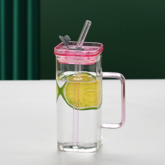 Clear glass mug with pink lid and handle, filled with a green drink and lime slice, on a white surface with green curtain background.