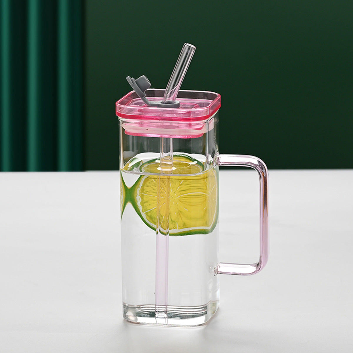Clear glass mug with pink lid and handle, filled with a green drink and lime slice, on a white surface with green curtain background.
