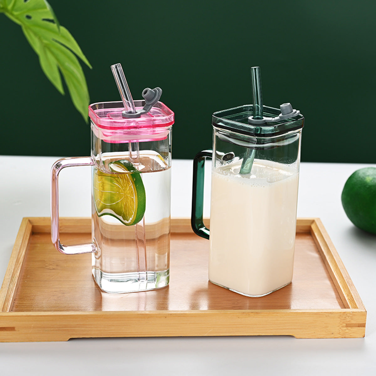 Two mugs with colorful lids and straws on a wooden tray with a green leafy background.